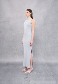 Vestido largo gris ajustado con cuello alto y aberturas laterales, hecho de una tela suave. Combinado con sandalias de tacón blancas sobre un fondo neutro.