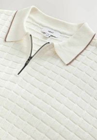 Polo shirt tricoté blanc texturé avec fermeture éclair partielle et bordure marron sur le col, posé à plat sur une surface claire.