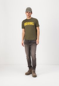 Olive green cotton t-shirt with yellow dripping ‘DIESEL’ logo, paired with grey jeans and beige lace-up boots. Hat in light grey with embroidered detail.
