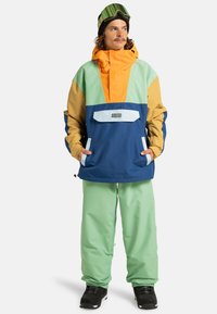 Mehrfarbige Skijacke mit orangefarbenen, grünen, marineblauen und tanfarbenen Abschnitten, ausgestattet mit einer Fronttasche und einem Reißverschluss. Dazu passende hellgrüne Hose.