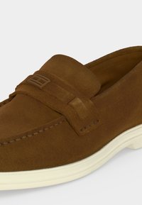 Καφέ μοκασίνι από σουέντ με ραφές μοκασίνου, λεπτομέρεια με λουράκι στην κορυφή και λευκή ελαστική σόλα.