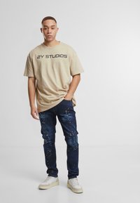 Béžové bavlněné tričko s potiskem "2Y STUDIOS" v černé barvě, spojené s tmavě modrými džíny s barevnými skvrnitými akcenty a bílými teniskami.