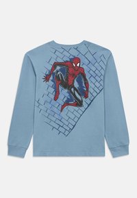 Dlhé rukávy modré tričko s grafickou potlačou červeno-čierneho Spider-Mana na chrbte na pozadí dizajnu tehlovej steny. Mäkká bavlnená látka.