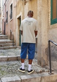 Lys beige oversized t-shirt med grafisk print på ryggen, parret med blå denimshorts og sorte sneakers med hvide såler og sokker.