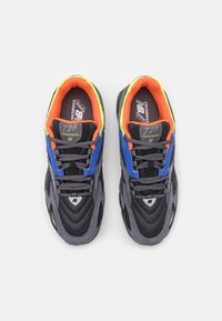 Harmaa ja musta New Balance 725 juoksukengät sinisillä yksityiskohdilla ja oranssin keltaisella sisävuorilla, nähtynä ylhäältä valkoista taustaa vasten.