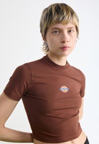 Top scurt maro cu maneci scurte, din material elastic, având un logo circular cu "Dickies" în albastru pe piept. Textură netedă.