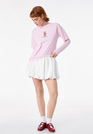 Junge Person, die ein pinkfarbenes lang- und kurzärmeliges Oberteil mit Teddybär-Motiv, einen weißen, aufgeplusterten Minirock, weiße Socken und rote Schuhe trägt.