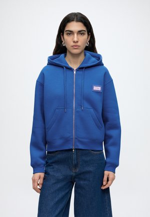 Femme aux longs cheveux noirs portant un sweat à capuche bleu zippé avec poches avant et étiquette sur la poitrine, associé à un jean bleu foncé, sur fond uni.