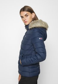 Chaqueta de plumas en color marino con capucha forrada de piel sintética, textura acolchada, bolsillos laterales y acento de logo en la manga. Cierre frontal con cremallera.