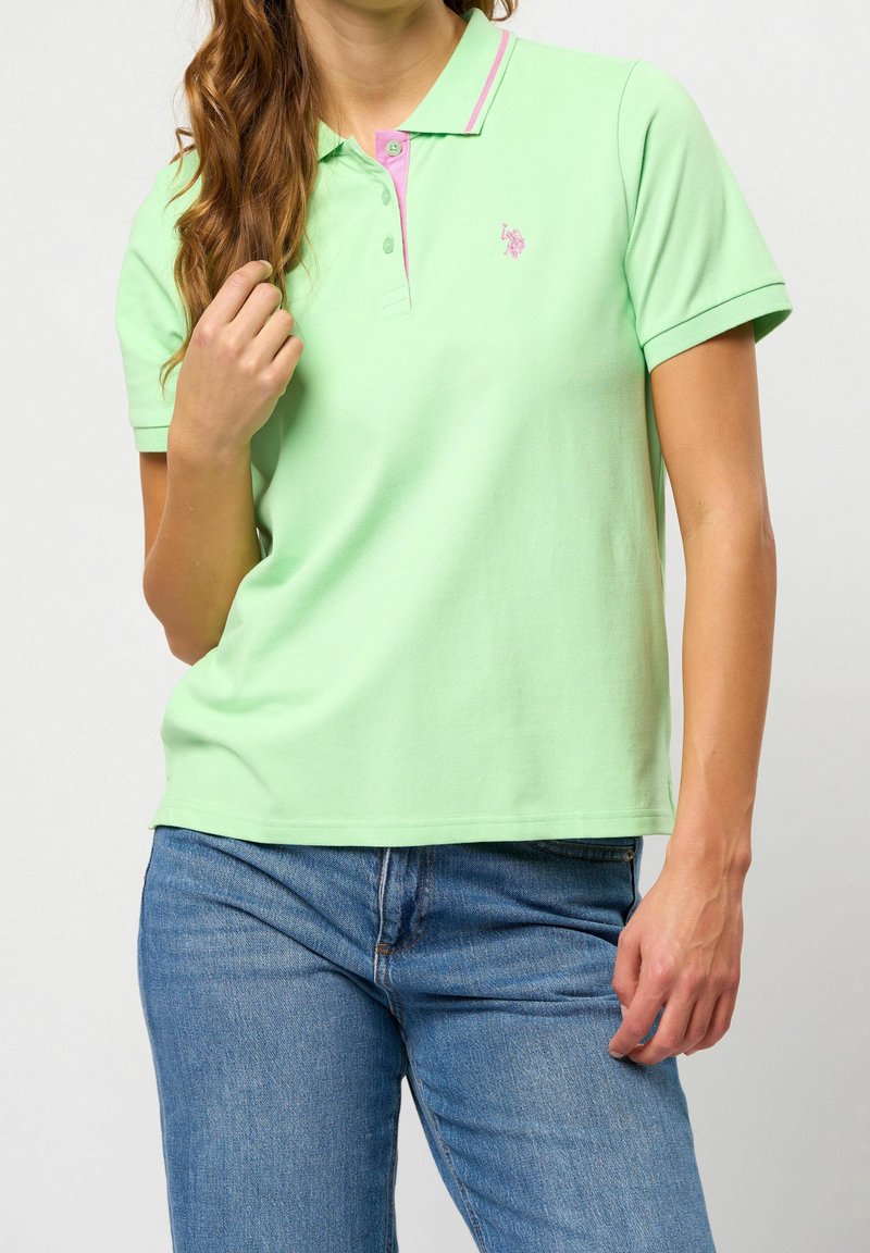 Femme portant un polo manches courtes vert clair avec des détails roses et un jean bleu, debout avec une main près du col.
