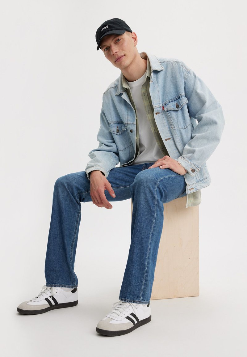 Helles blauer Jeansjacke, graues T-Shirt, olivgrüne Überhemd, blaue Jeans und weiße Turnschuhe mit schwarzen Streifen, auf einem Holzblock sitzend.