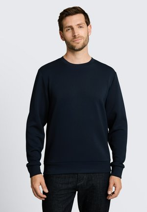 Mann trägt ein schlichtes dunkelblaues Sweatshirt mit langen Ärmeln und dunkle Jeans, steht vor einem hellgrauen Hintergrund.