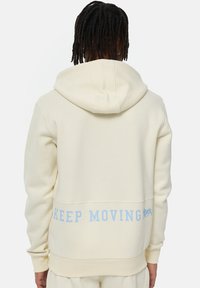 Krémová mikina vyrobená z mäkkej látky, s klokankovou vreckom a modrým nápisom "KEEP MOVING" na zadnej strane. Obsahuje kapucňu so sťahovacou šnúrkou.