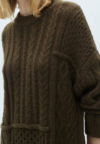 Pull en tricot marron à col rond, présentant un motif texturé et une poche avant. Le tissu en tricot doux et épais apporte de la chaleur.