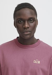 Mørkerød sweatshirt med rund hals, der har et lille "KEEP CAℓM" grafik i beige. Glat stoftekstur, afslappet pasform.