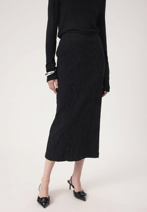 Kobieta ubrana w długą, czarną spódnicę o fakturze, czarną bluzkę z długim rękawem, srebrną bransoletkę oraz czarne, szpiczaste buty na wysokim obcasie typu slingback.