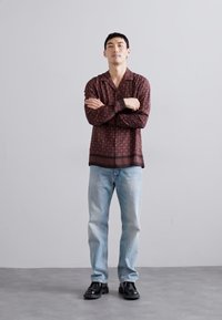 Langærmet bordeauxmønstret skjorte, lyseblå jeans og sorte sko. Skjorten har en button-down krave og en horisontal kantdetalje.