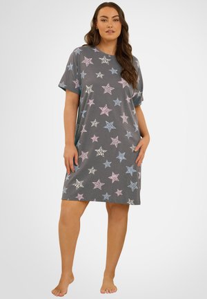 Vrouw die een losse grijze jurk tot op de knie draagt met roze, blauwe en witte sterpatronen, blootsvoets staand tegen een effen achtergrond.