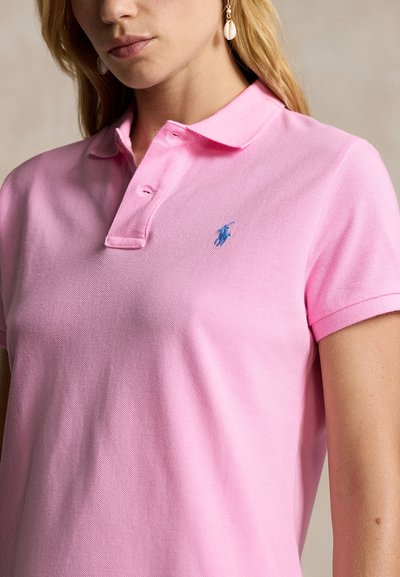 Woman wearing a light pink polo shirt with a small blue embroidered logo on the left chest and seashell drop earrings.