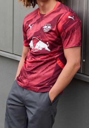 Person iført en rød og mørkerød mønstret sportstrøje med hvide Puma- og Red Bull-logoer, stående med hænderne i lommerne på grå bukser.