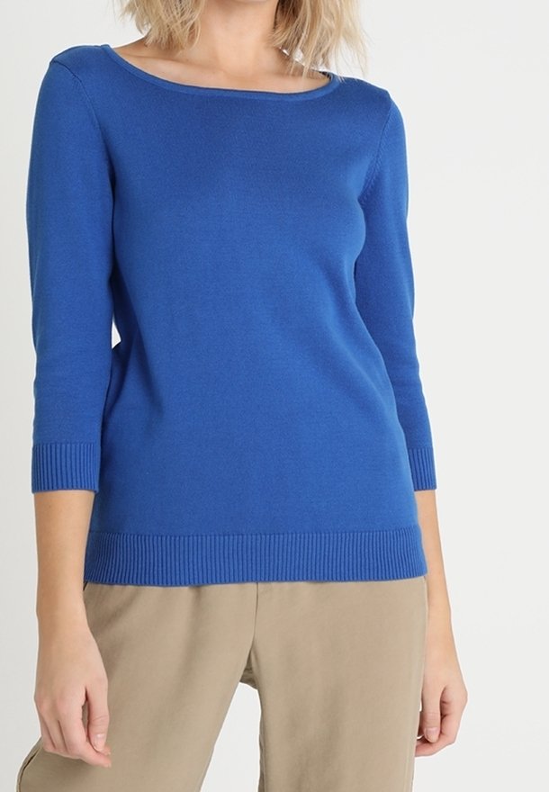 Femme portant un pull bleu ajusté à manches trois-quarts et un pantalon beige sur un arrière-plan clair uni.