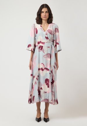 Žena oblečená v svetlomodrých zavinovacích šatách s ružovým a bordovým kvetinovým vzorom a 3/4 naberanými rukávmi, v kombinácii s čiernymi lodičkami so špicatou špičkou.