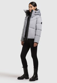 Giacca imbottita grigia chiara con colletto alto, chiusura con zip e cappuccio regolabile. Presenta un design trapuntato e dettagli neri. Abbinata a leggings neri e stivali.