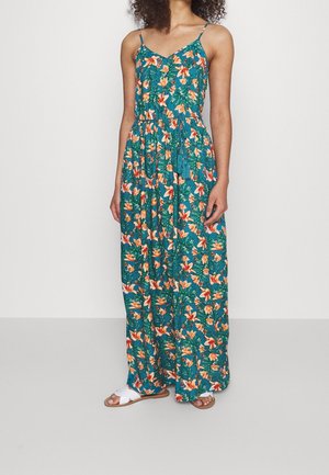 Frau trägt ein Teal Maxi-Kleid mit orangefarbenem Blumenmuster, Spaghettiträgern und einem Taillendetail, kombiniert mit weißen flachen Sandalen.