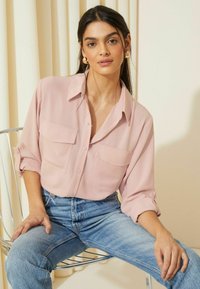 Hellrosa Button-Up-Hemd mit zwei Brusttaschen, hochgekrempelten Ärmeln und einem lockeren Schnitt, kombiniert mit hochgeschnittenen blauen Jeans.