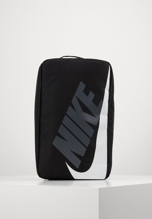 Sac rectangulaire noir Nike avec grand texte gris Nike et logo swoosh blanc à l'avant, debout sur une surface blanche.