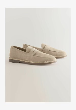 Beige suède instappers met een gestikte rand, een platte zool en een band over de bovenkant. Eenvoudig, minimalistisch ontwerp.