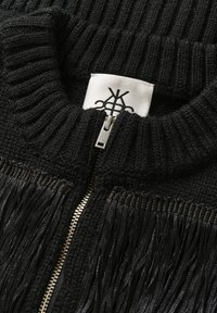 Nahaufnahme eines schwarzen gestrickten Kleidungsstücks mit silbernem Reißverschluss und schwarzen Fransenverzierungen, die ein weißes Stoffetikett mit einem schwarzen abstrakten Logo zeigen.