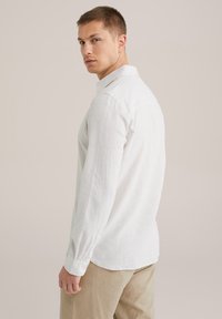 Camisa blanca de manga larga con tejido texturizado, puños abotonados y cuello clásico, llevada con pantalones beige claros, vista desde la parte de atrás.