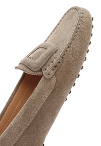 Mocassino in suede beige con una tracolla decorativa, dettagli ricamati e suola in gomma chiodata per aderenza e flessibilità.