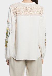 Kvinna som bär en krämfärgad långärmad blus med spetsaxelparti och blommig broderi på ärmarna, i kombination med beiga byxor, sedd bakifrån.