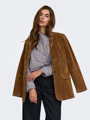 Mujer con una chaqueta de ante marrón sobre una camisa de cuadros azul y blanca con pantalones negros, de pie frente a un fondo claro y liso.