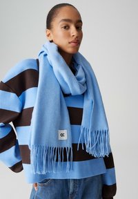 Écharpe bleu clair en tissu doux avec bords frangés, portée sur un pull rayé bleu et marron. Étiquette visible sur l'écharpe.