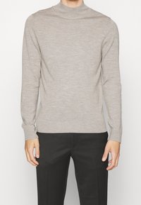 Homme portant un pull léger beige ajusté à manches longues et un pantalon noir, se tenant devant un fond blanc uni.