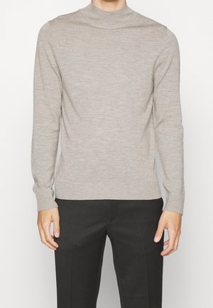 Mann in einem taillierten, hellbeigen Langarmpullover und schwarzen Hosen, stehend vor einem schlichten weißen Hintergrund.