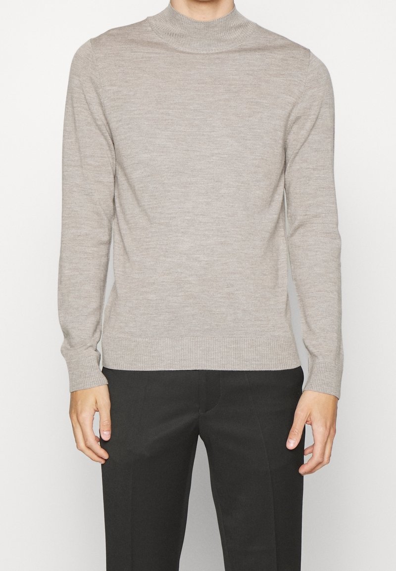 Homme portant un pull léger beige ajusté à manches longues et un pantalon noir, se tenant devant un fond blanc uni.