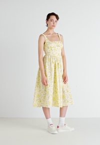 Žlté kvetinové midi šaty so špagetovými ramienkami, nariaseným živôtikom a rozšírenou sukňou, kombinované s bielymi teniskami a ponožkami po členky.