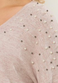 Vaaleanpunainen neule, jossa on V-pääntie ja ripoteltuja helmiä sekä metallipainotteisia koristeita, valmistettu pehmeästä ja teksturoidusta kankaasta.