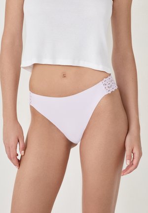 Donna che indossa uno slip bikini di un leggero colore viola con dettagli in pizzo floreale sui fianchi e un top corto bianco, in piedi contro uno sfondo semplice.