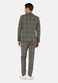 Mann steht mit dem Rücken zugewandt und trägt einen grauen karierten Anzug mit Jacke und Hose, kombiniert mit weißen Sneakers vor hellem Hintergrund.