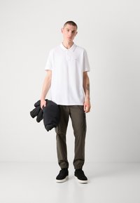 Hvit polo skjorte med en liten logo, sammen med olivengrønne cargo bukser. Holder en svart boblejakke. Sorte joggesko med hvit såle.