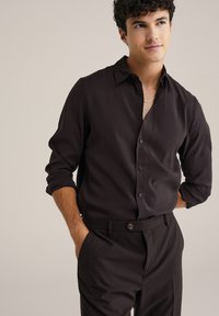 Črna majica z dolgimi rokavi in gumbnim zapenjanjem ter ovratnikom, izdelana iz gladkega blaga, nekoliko oprijetega kroja, v kombinaciji s temnimi hlačami.