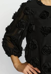 Sort kjole med trekvartlange ærmer, pyntet med 3D-stofroser. Gennemsigtig tekstur på ærmerne med elastiske manchetter og en enkel halsudskæring.