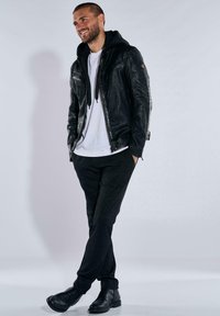Schwarze Lederjacke mit Kapuze über einem weißen T-Shirt, kombiniert mit dunklen Hosen und schwarzen Schuhen. Die Jacke hat Reißverschlüsse und strukturierte Details.