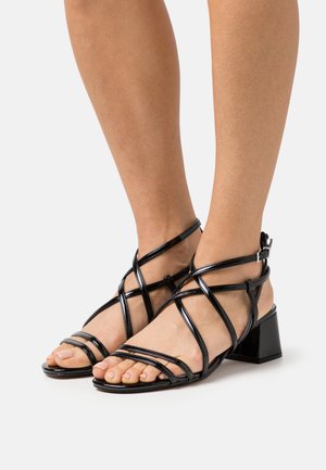 Sandal cu toc bloc, din piele patent neagră, cu bretele crisscross și închidere cu cataramă. Design cu vârful deschis, cu o finisare netedă și lucioasă.