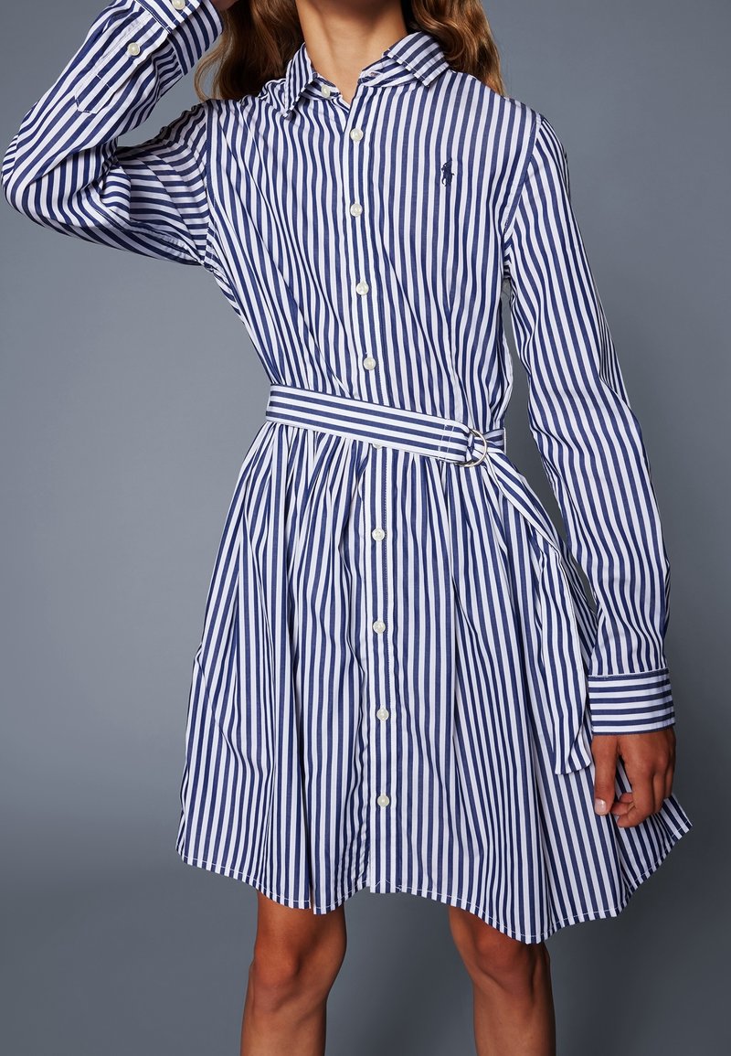 Mladý člověk oblečený v modro-bíle pruhované košilové šaty s odpovídajícím páskem, stojící proti jednoduchému šedému pozadí.